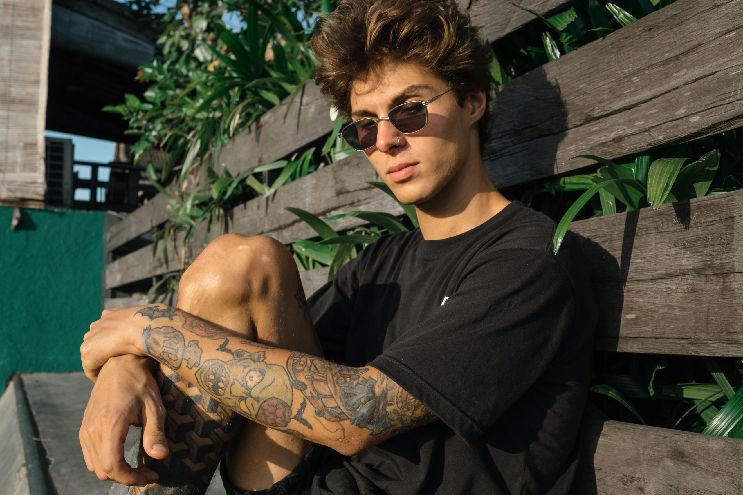 Teenager with tattoos and sunglasses sitting on a bench in casual attire, enjoying the sun.